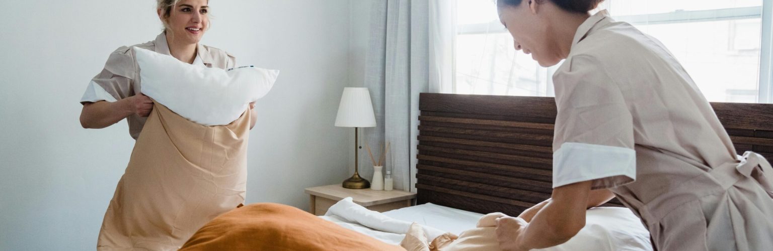 Two women in uniforms arranging bed linens in a clean and bright bedroom.